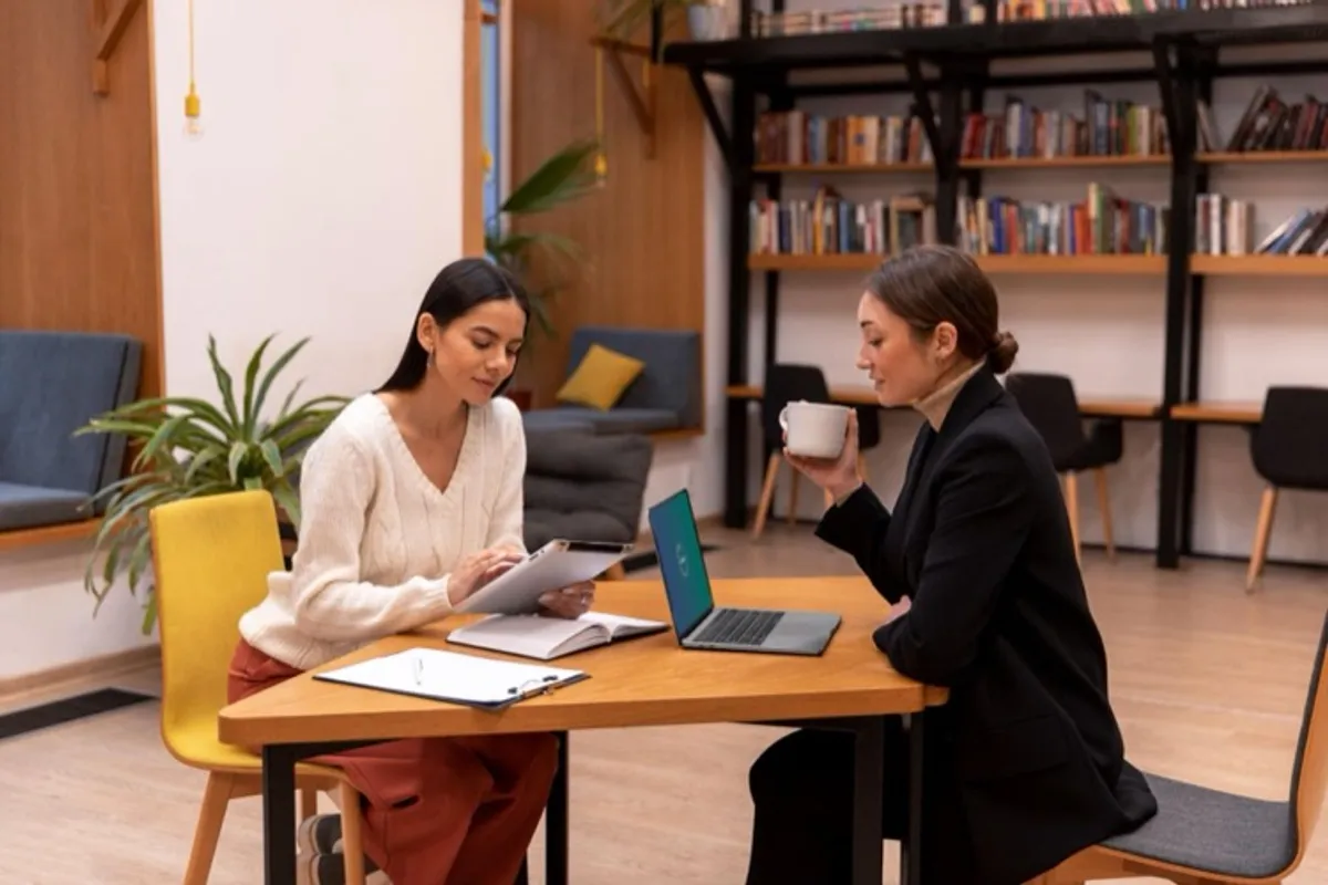 Two women sitting at a table in a library-like setting, one holding a tablet and the other with a cup of coffee and a laptop, engaged in a discussion